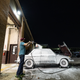 nighttime photo of the 1974 bmw 2002tii parked outside a large garage. the car is covered top to bottom in soap suds as the owner sprays the car with a pressure washer.